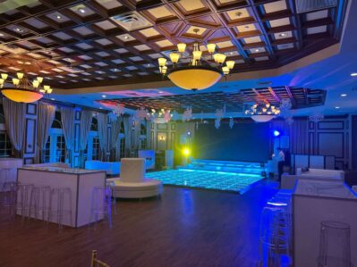 Elegant event hall in NY with a glowing LED dance floor, modern white seating, transparent chairs, stylish chandeliers, and large windows with drapes. Blue and yellow lighting plus DJ services Suffolk and Nassau County, NY create a festive atmosphere.