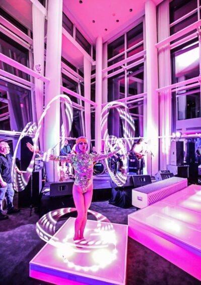 A performer in a colorful bodysuit hula hoops with glowing LED hoops on a lit platform at an NY,NY indoor event, surrounded by large windows and pink lighting, with a band playing—perfect for entertainment services in Suffolk and Nassau County.