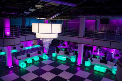 Modern NY event space with a black and white checkered dance floor, large chandelier, glowing green and purple LED-lit furniture, white sofas, round tables, and purple ambient lighting—perfect for parties with DJ services in Suffolk and Nassau County, NY.