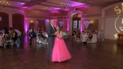 A young woman in a bright pink gown dances with an older man at a formal event, possibly a quinceañera, in a ballroom bathed in pink lighting. Guests look on as top DJ services Suffolk and Nassau County, NY set the perfect mood for the celebration.