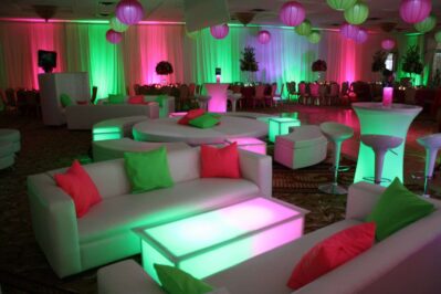 A modern event space with white furniture, neon pink and green lighting, glowing tables, colorful throw pillows, round banquet tables, and hanging paper lanterns—perfect for vibrant parties and entertainment services in Suffolk and Nassau County, NY.