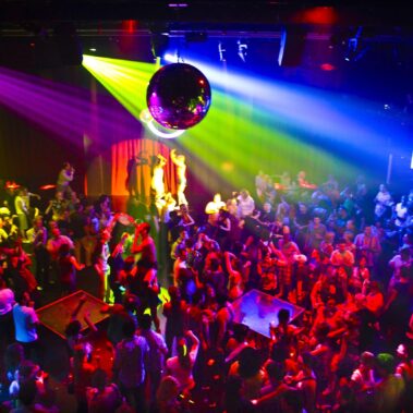 A crowded nightclub dance floor filled with people under vibrant, colorful lights, a disco ball overhead, and a DJ on an elevated booth—perfect for those seeking top entertainment services in Suffolk and Nassau County, NY.