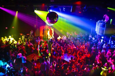 A crowded nightclub dance floor filled with people under vibrant, colorful lights, a disco ball overhead, and a DJ on an elevated booth—perfect for those seeking top entertainment services in Suffolk and Nassau County, NY.