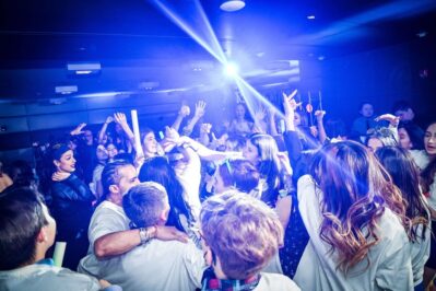 A large group of young people dance and celebrate under blue lights in a crowded NY,NY nightclub, with arms raised and smiles on their faces, creating a lively and energetic atmosphere enhanced by top DJ services Suffolk and Nassau County.