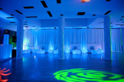 A modern, empty event space in NY with blue lighting, shiny pillars, draped white curtains, and white furniture. Green and blue light patterns dance on the wooden floor. Perfect for entertainment services in Suffolk and Nassau County, the atmosphere is sleek and elegant.