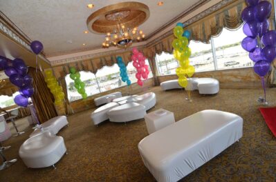 A decorated event space with white modern seating, colorful balloon clusters, large windows, gold accents, chandeliers, and patterned carpet—perfect for entertainment services in Suffolk and Nassau County, NY.