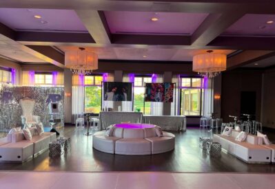 A modern event space with white circular seating, silver décor, and chandeliers. Purple lighting accents the room, and large windows let in natural light. Perfect for entertainment services Suffolk and Nassau County, NY. Two large art pieces hang above a decorated table.