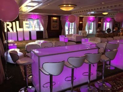 A stylish event space with modern white furniture, pink lighting, and illuminated "RILEY" letters. Perfect for celebrations, it offers a festive vibe and access to premier entertainment services Suffolk and Nassau County, NY.