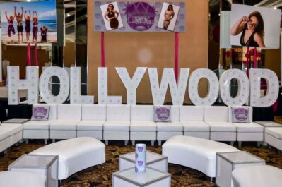 Large white marquee letters spell "HOLLYWOOD" behind a white lounge seating area, perfect for NY events. Posters featuring women in various poses hang above, adding to the glamorous, modern party vibe ideal for entertainment services Suffolk and Nassau County.