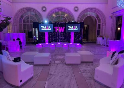Luxurious event space with white seating, purple lighting, and a stage featuring two screens displaying “TALIA” and a large “T W.” Elegant arched windows frame the background—perfect for events with top DJ services in Suffolk and Nassau County, NY.