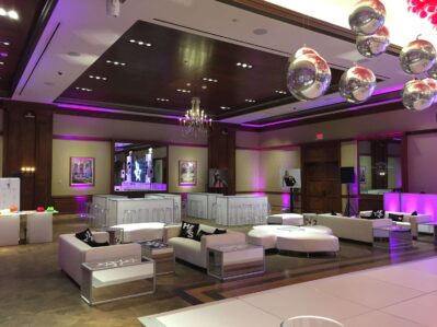 A modern NY event space with white lounge furniture, purple accent lighting, mirrored disco balls, and framed art. The room features a large wooden door, chandeliers, and a bar—perfect for entertainment services in Suffolk and Nassau County.