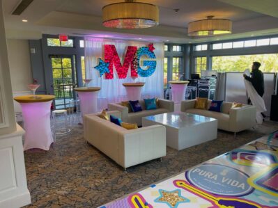 A decorated NY event space features white couches, pink-lit cocktail tables, and a large “MG” sign with stars. A person arranges linens as the floor displays vibrant graphics and a “Pura Vida” message—perfect for entertainment services Suffolk and Nassau County.