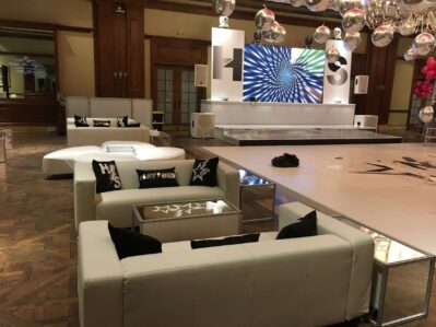 A modern NY event space features white couches, mirrored tables, a stage with a digital spiral display, silver decor, and hanging disco balls. Pink balloons add color—perfect for entertainment services Suffolk and Nassau County.