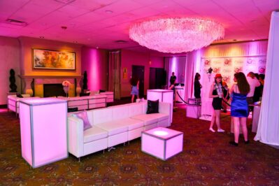 A modern lounge with white couches and glowing pink accent lighting. Several people in semi-formal attire mingle near a photo backdrop. DJ services Suffolk and Nassau County, NY enhance the vibe while art decorates the walls beneath a large chandelier.