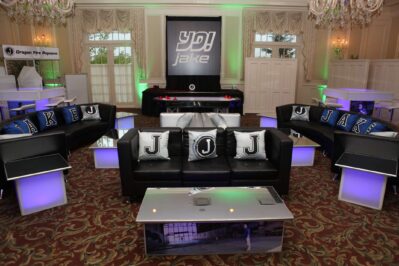 A lounge area with black couches and "JAKE" pillows, illuminated tables, and a large "YO! Jake" sign in an event space with chandeliers—perfect for NY events seeking top entertainment services in Suffolk and Nassau County.