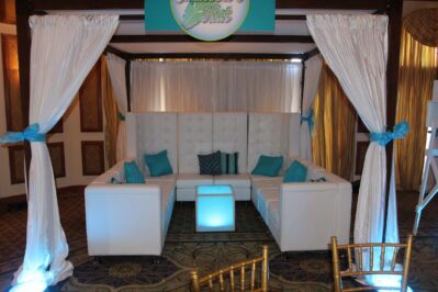 A white lounge seating area with teal accent pillows and a glowing square table, framed by white curtains tied with teal ribbons, in an elegant NY room offering entertainment services in Suffolk and Nassau County.