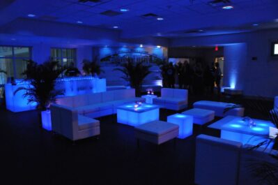 A modern lounge with white couches, ottomans, and tables illuminated by blue LED lighting; several people gather near the bar in the background under a dim, ambient atmosphere—ideal for events needing DJ services in Suffolk and Nassau County, NY.