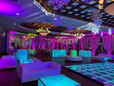 A large event hall in NY decorated with glowing purple and blue lights, modern white furniture, illuminated tables and dance floor, hanging snowflake decorations from the ceiling, and top dj services Suffolk and Nassau County, NY.