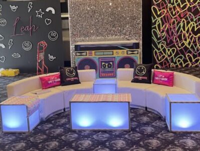 A curved white couch with glowing side tables, colorful pillows, and a bold backdrop featuring a large boombox, graffiti hearts, and neon doodles—perfect for NY events or dj services in Suffolk and Nassau County, NY. "Leah" shines on the chalkboard wall.