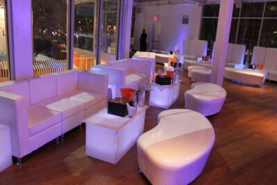 A modern lounge with white sofas, curved benches, illuminated tables, and orange ice buckets on wood floors, set in a glass-walled room offering city lights—the perfect venue for dj services Suffolk and Nassau County.
