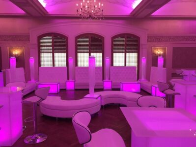 A modern lounge with white, futuristic furniture, high-backed seats, and stools illuminated with pink-purple LED lighting—perfect for events in Suffolk and Nassau County, NY—set in an elegant room with chandeliers and arched windows.