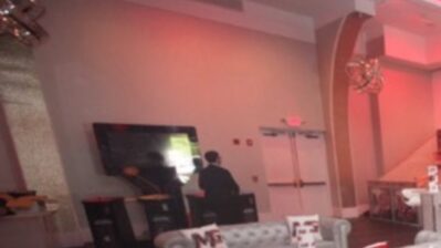 A person stands near a large screen in a modern, softly lit NY room with white walls, double doors, decorative pillows on a couch, and unique lighting fixtures. The overall ambiance has a warm, reddish tint.