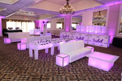 A stylish event lounge with white modern furniture, glowing purple accent lights, a foosball table, and a bar area with "Isabel" signs—plus top entertainment services Suffolk and Nassau County—all under elegant chandeliers in a spacious banquet hall.