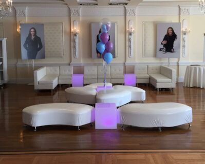 A modern lounge area in NY with white curved seating, purple-lit cube tables, and a cluster of purple and blue balloons—perfect for entertainment services Suffolk and Nassau County. Elegant cream walls feature large portraits of a young woman.