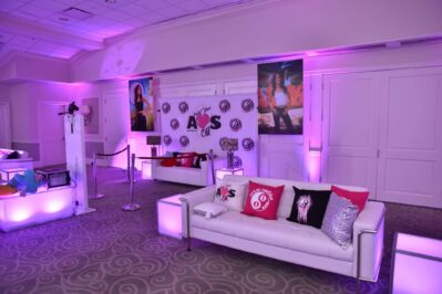 A lounge area with white sofas, colorful pillows, and purple lighting. Large posters and a decorated backdrop with “AKS” create a festive vibe—perfect for entertainment services Suffolk and Nassau County, NY.