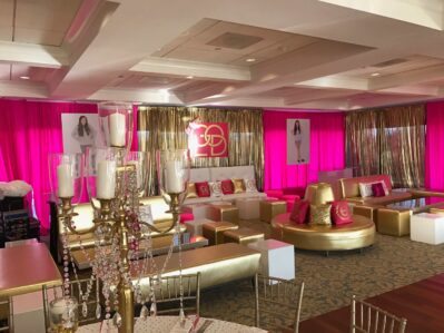 A glamorous event space with hot pink and gold decor, white and gold seating, gold drapes, and framed photos of a young woman — perfect for entertainment services Suffolk and Nassau County, NY. A chandelier with crystal accents graces the foreground.