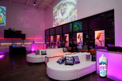 A modern NY event space with white sofas, colorful “Rock the Bells” pillows, and posters of women on glass walls, illuminated by pink and purple lights. Entertainment services Suffolk and Nassau County are available for any occasion.