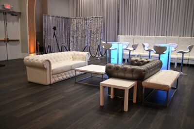 Modern lounge area in NY, NY, featuring white and gray tufted sofas, glowing lighted tables, barstools, and silver curtains. Perfect for entertainment services Suffolk and Nassau County. Dark wood flooring adds a stylish touch.