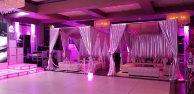 A luxurious NY event venue with white tufted sofas, canopy drapes, pink-purple lighting, a dance floor, and chandeliers—perfect for hosting unforgettable moments with top entertainment services in Suffolk and Nassau County.
