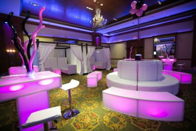 A modern event space with glowing pink and purple LED furniture, white curved seating, high chairs, draped cabanas, and balloon decorations under elegant chandeliers—perfect for entertainment services in Suffolk and Nassau County.