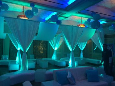 A lounge area in NY, NY, decorated with white drapes, sofas, and glowing blue lights creates an elegant vibe. Blue paper lanterns hang from the ceiling while one person stands near the right—perfect for entertainment services Suffolk and Nassau County.