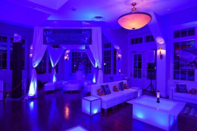 A modern lounge in NY, decorated with white furniture and draped fabric under blue lighting, featuring a “Jesse’s Grad Lab” banner, glass doors, large windows, and a glowing square table—perfect for entertainment services Suffolk and Nassau County.