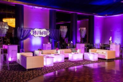 Elegant event space with purple lighting, modern white sofas, glowing tables, chandeliers, and a sign reading “HANNAH.” Perfect for any celebration, this stylish venue also offers top DJ services in Suffolk and Nassau County.