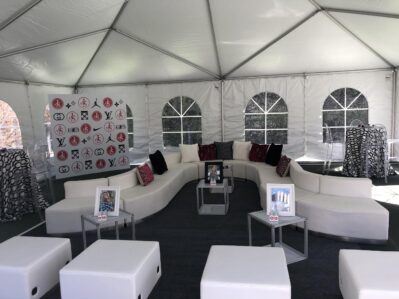 A white canopy tent with curved white sofas, small tables holding framed photos, and decorative pillows. Perfect for NY, NY events or entertainment services in Suffolk and Nassau County. A logo backdrop and arched windows complete the inviting space.