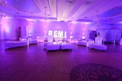 A spacious event hall with purple lighting, white lounge furniture in a semi-circle, and large illuminated "REMI" letters at the center. Two people stand by a shimmering silver backdrop—perfect for DJ services Suffolk and Nassau County, NY.