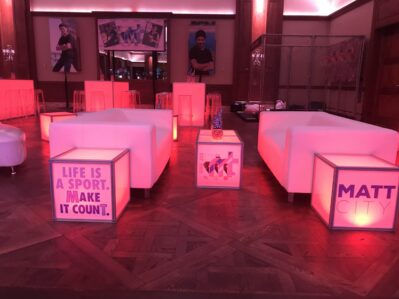 A modern NY lounge with glowing pink cube tables, white sofas, and motivational signs like "LIFE IS A SPORT. MAKE IT COUNT." and "MATT CITY." A person stands behind a bar—perfect for events needing DJ services in Suffolk and Nassau County, NY.