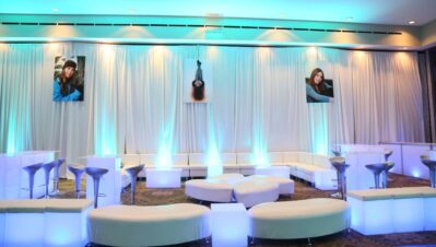 A modern NY,NY lounge with white curved sofas, glowing blue-lit tables, silver stools, and draped walls decorated with three large photos of a young woman. Soft blue lighting and dj services Suffolk and Nassau County enhance the ambiance.