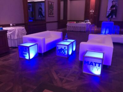 A lounge area in NY features white couches and glowing blue cube tables with phrases like “MATT CITY” and “LIFE IS SHORT. MAKE IT COUNT.” Ideal for entertainment services Suffolk and Nassau County, buffet tables grace the stylish parquet floor backdrop.
