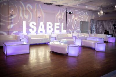 A modern lounge with white furniture and glowing tables, featuring large illuminated “ISABEL” letters against a light-filled wall. Perfect for events with professional DJ services in Suffolk and Nassau County, NY. Chandeliers and wooden flooring complete the scene.