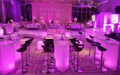 Elegant NY event space with modern white furniture, high tables with barstools, banquet tables, and soft pink-purple ambient lighting. Crystal decorations and floral centerpieces add a luxurious touch—perfect for entertainment services Suffolk and Nassau County.