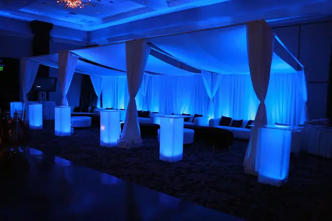 A modern indoor event space with white draped fabric, illuminated by blue lighting. There are glowing pillars, white sofas, and elegant decor, creating a sophisticated and stylish atmosphere.