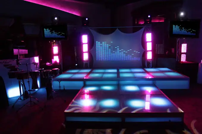 A brightly lit dance floor with glowing blue and pink squares, surrounded by colorful LED lights, speakers, and musical instruments, set up for a lively party or event.