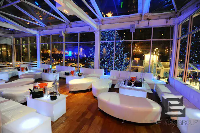 A modern rooftop lounge with white sofas and tables, wooden floors, floor to ceiling windows, and blue accent lighting. A large decorated tree with blue lights is visible outside, along with city buildings at night.