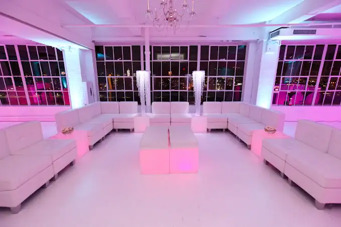 A modern lounge with white sofas arranged in a U shape, illuminated by pink and purple lighting. Large windows reveal a nighttime city view, and two tall decorative light fixtures stand inside the room.