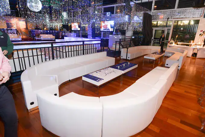 A modern lounge with white curved sofas, low tables, and polished wooden floors. The room is decorated with hanging lights and has screens on the walls, creating an elegant, festive atmosphere.