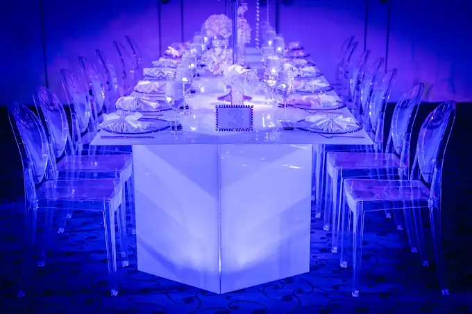 A long banquet table with white plates, napkins, and clear acrylic chairs, set for a formal event under dramatic blue lighting. White floral centerpieces and candles add elegance to the setting.